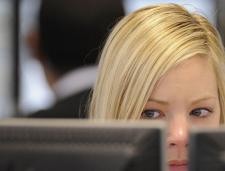 A blonde white woman looks suspiciously out of the corner of her eyes, onto a computer screen that is in front of her. 
