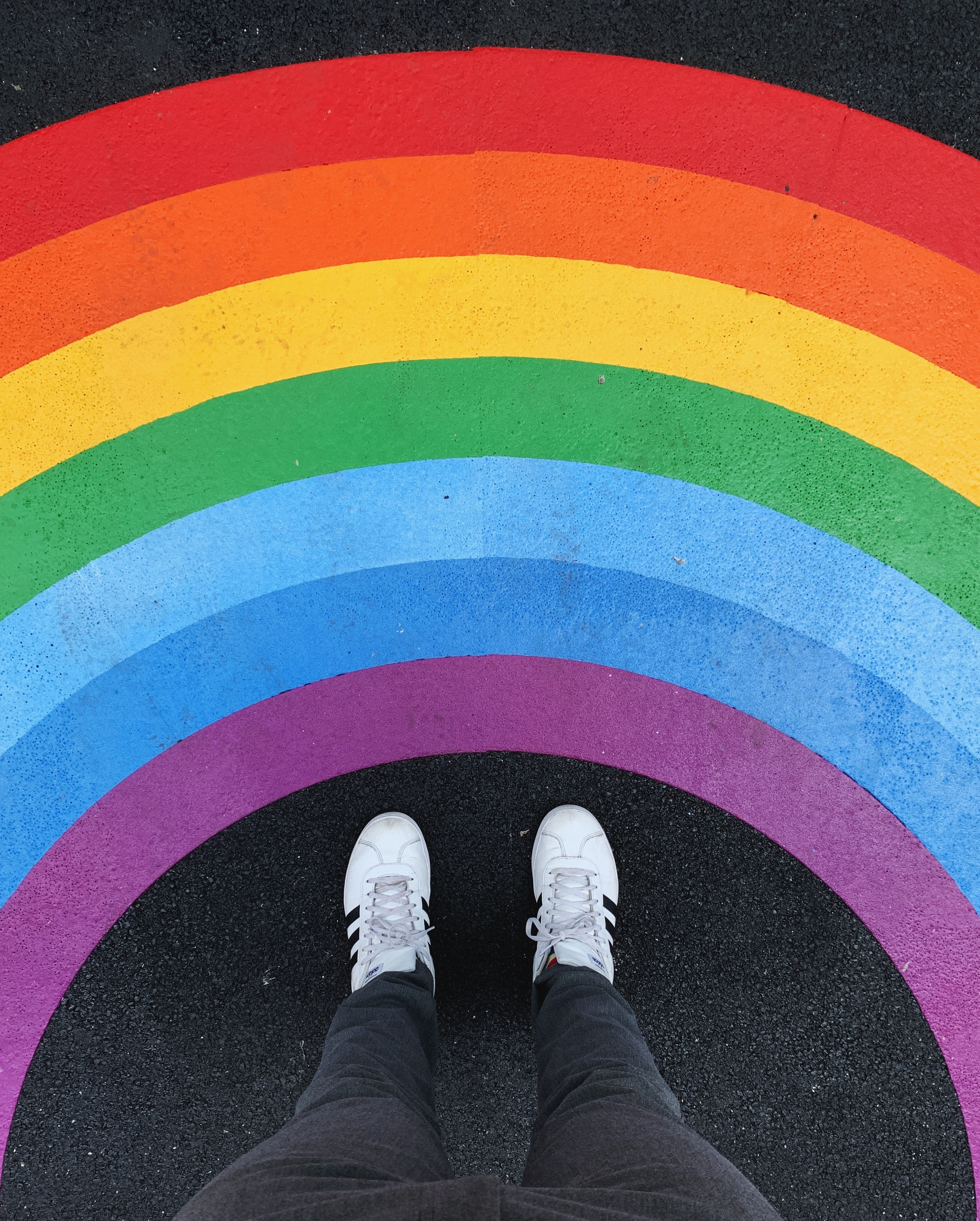 A vibrant rainbow is painted onto a road. Two feet wearing white shoes are situated at the bottom of the picture facing the rainbow