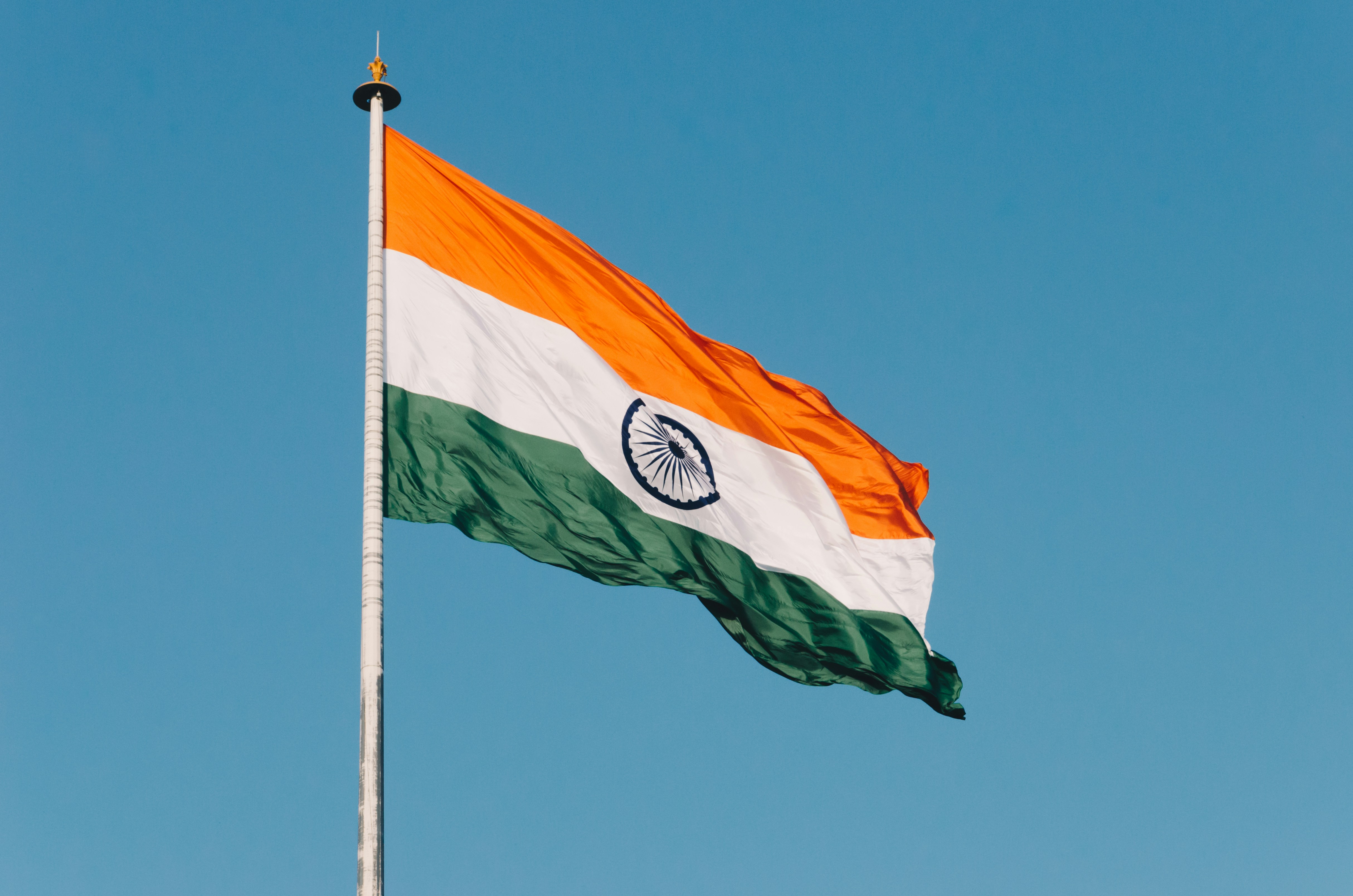 Image shows the Indian flag, comprised of 3 colored stripes of orange, white and green from top to bottom with an emblem at the center of the middle white stripe, flying high with the sky as the background.