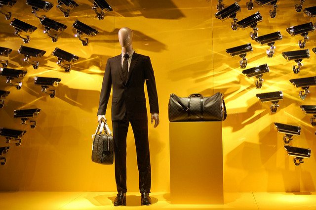 An art piece displaying a central mannequin figure in a work suit holding a luggage bag. Surrounding this figure are several surveillance cameras fixated on the figure.