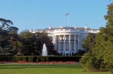Landscape photo of the US White House. The American flag flies above the top and a water fountain flows in the front of the house.Trees are surrounding. 