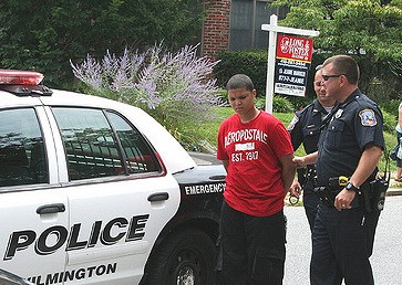 Two white officers handcuff a young black teen on the side of the police vehicle.