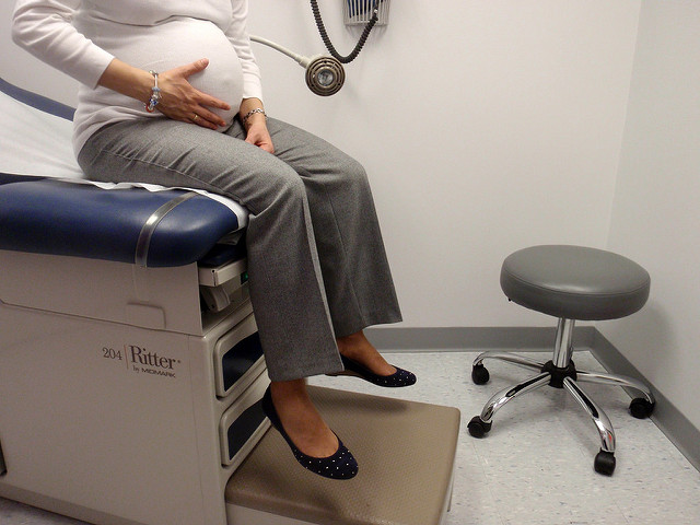 A pregnant woman sits on patient bed in a doctor's office, with right hand caressing stomach. 