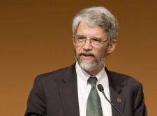 John Holdren is pictured against a solid mustard yellow background.