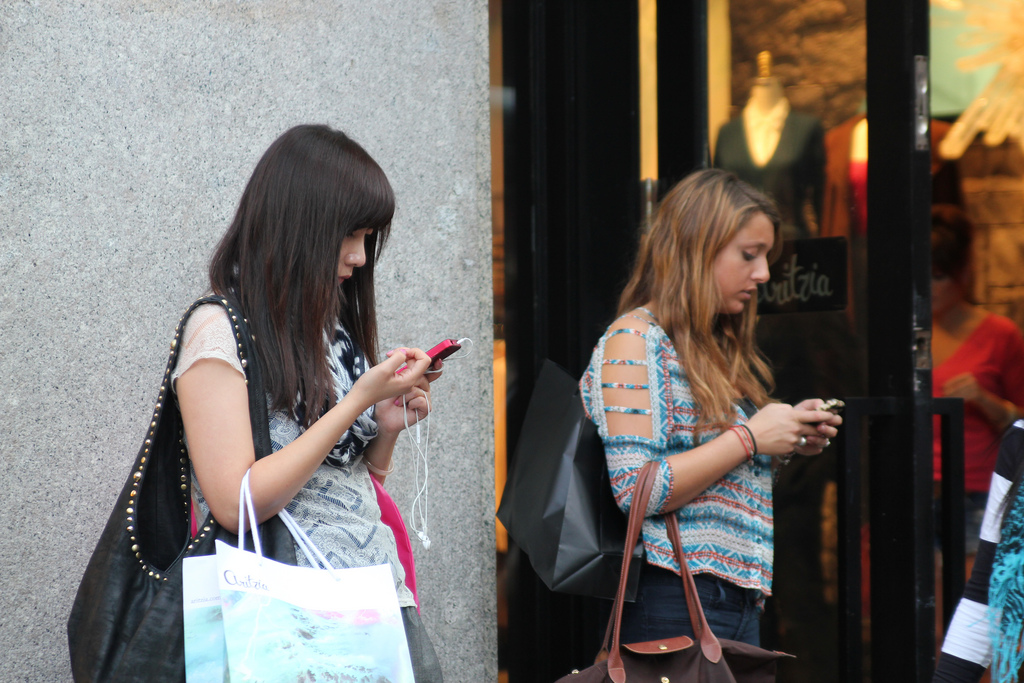 Two young women of color carry shopping bags on their shoulders, and look down at their phone in their hands.