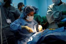 A surgeon holding instruments is surrounded by a team. A light beams on patient undergoing surgery.