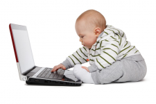 A baby sits in front and engages with a laptop.