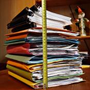 A measuring tape lines a stack of folders and bound books on a table.
