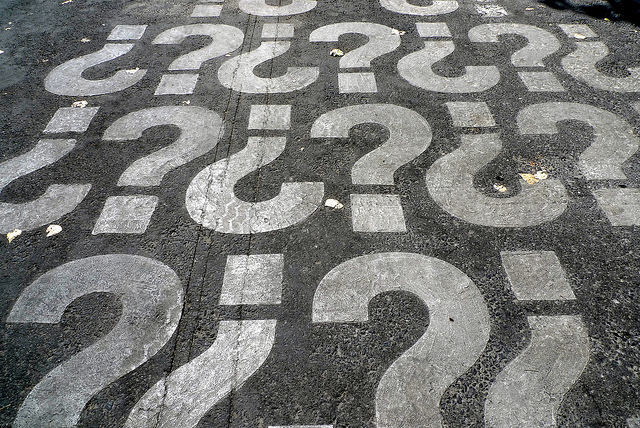 Repeated pattern of question marks, on asphalt surface. 