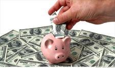 A piggy bank is surrounded by one hundred dollar bills. A hand is extended to place a bill into the piggy bank.