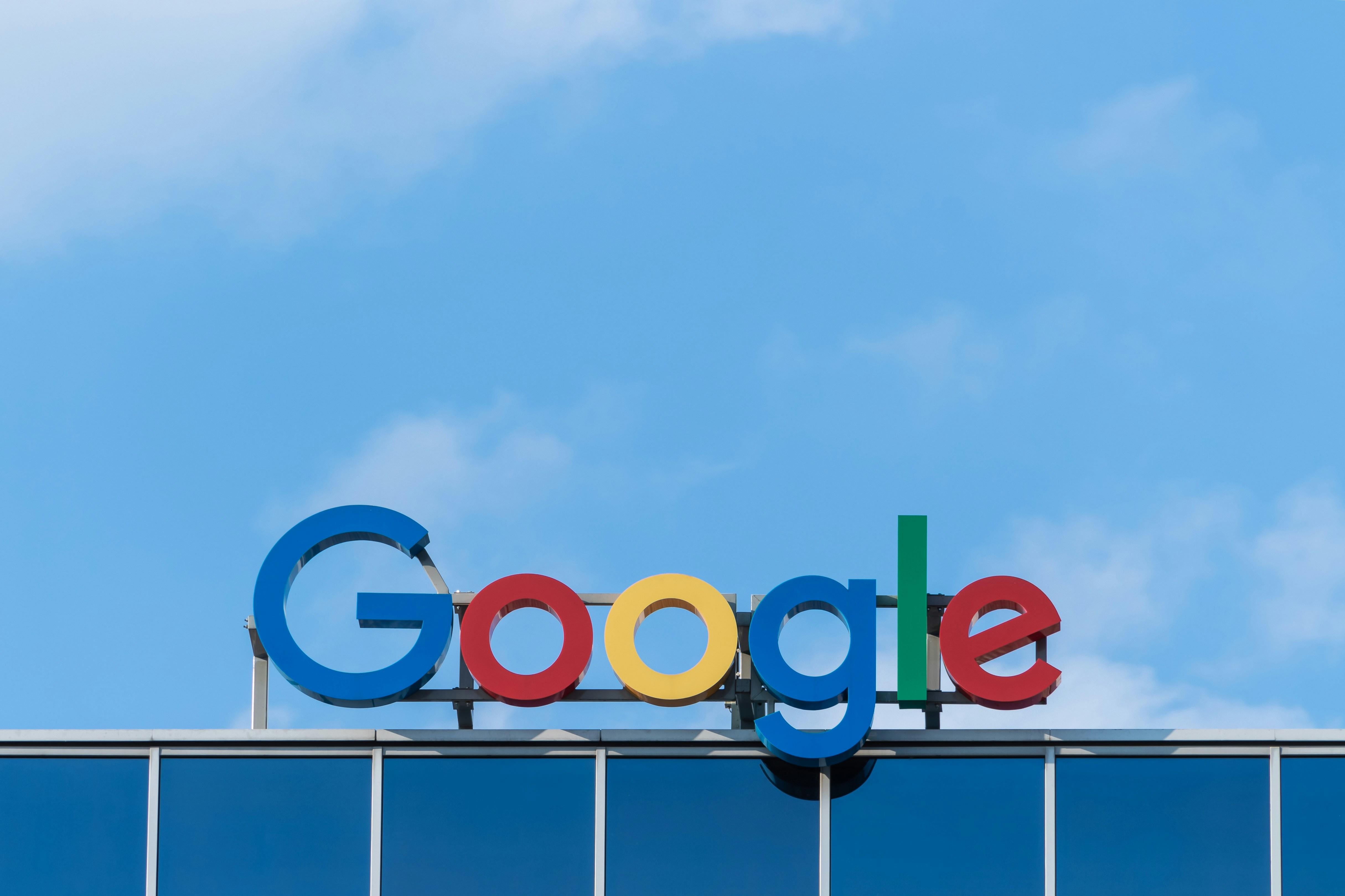 Image is the multi-colored sign of Google's logo at the top of a building, in which each letter alternates in color in the order of blue for G, red for o, yellow for o, blue for g, green for l, and red for e. 