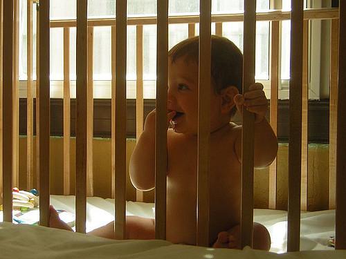 A baby, sitting inside of a crib, playfully looks to the side. One hand is holding a bar of the crib. Light pours out from a nearby window.