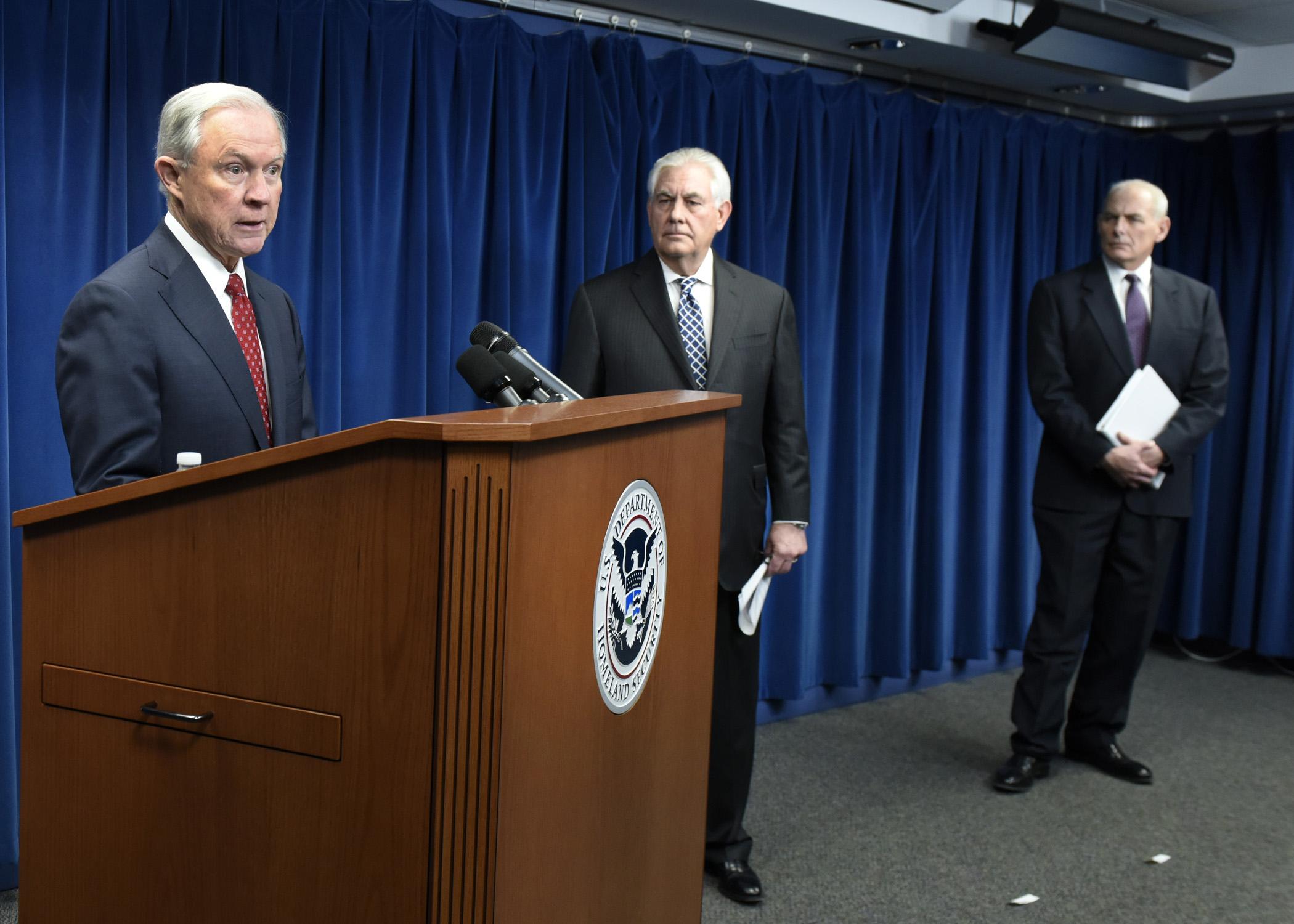 Attorney General Jeff Sessions stands behind podium at a press conference. Secretary of State Rex Tillerson and Secretary of Homeland Security John Kelly are looking towards him from the side.
