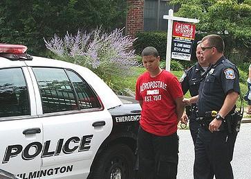 Two white officers handcuff a young black teen on the side of the police vehicle.