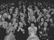 A black and white photo of an enthusiastically excited audience cheering, clapping, and smiling.