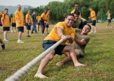 Two teammates of a tug-of-war game are pictured, with competitive looks on their faces as they cling to the rope. Onlookers look entertained.