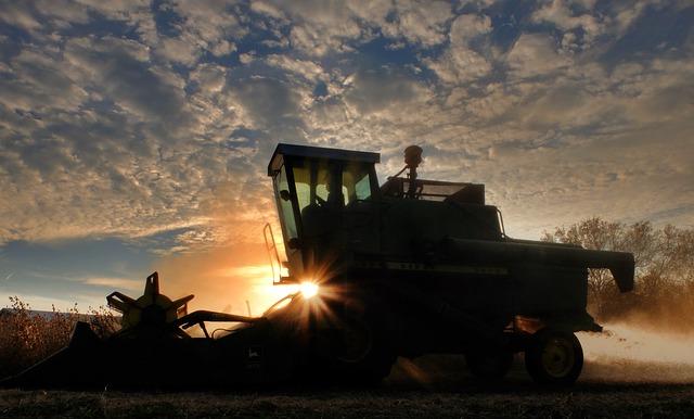 Silhouette of a tractor in a crop field. Against its background is a sunset, and blue skies with clouds
