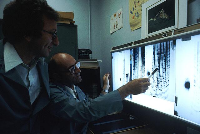  Two male scientists wearing lab coats in a laboratory looking at an illuminated board, reading the genetic code in the DNA.