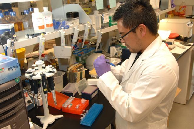 A scientist in a lab coat concentrates on writing on a test tube sample. He is surrounded by lab equipment, including pipettes, liquid solutions, and test tube racks.