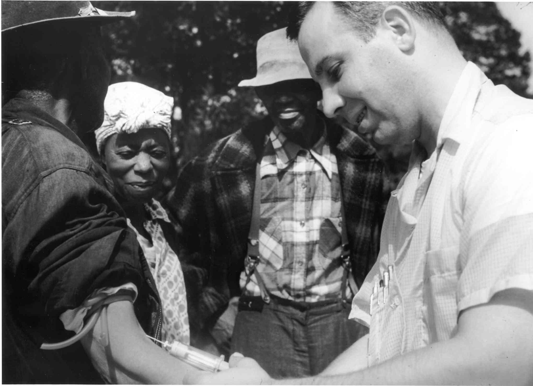 Archived black and white photo from the Tuskegee study. A white doctor draws blood from a black study participant with two others observing. 