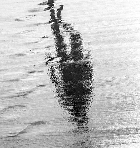 Grayscale photo of a person's refection from a puddle of water, as they walk .