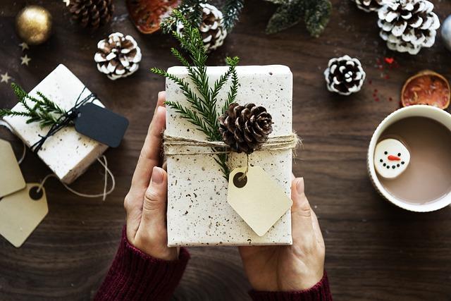 A wrapped gift is held by a person's hands. In the background are winter seasonal items, such as a mug filled with a beverage, pine cones, and another wrapped gift.