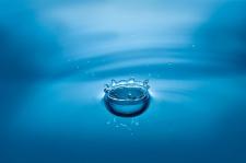 A water droplet falls into a larger pool of liquid, leaving behind ripples to the surface.