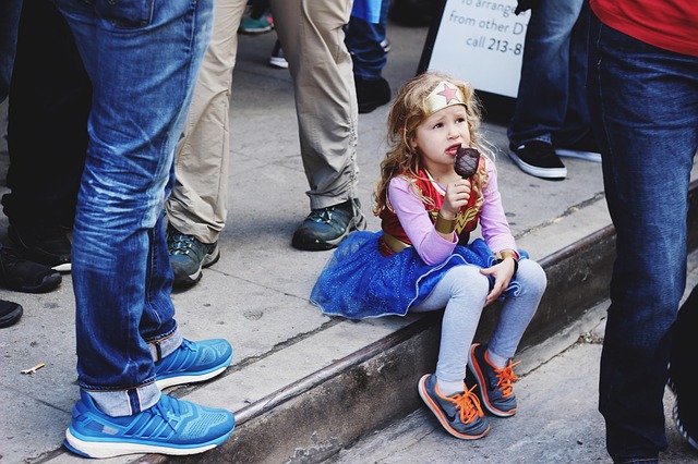 A little girl, dressed in a Wonder Woman's costume, sits on a sidewalk eating ice cream, while adults walk around her.